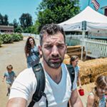 Man with a backpack takes a selfie on a sunny farm path, with kids and a white tent in the background.