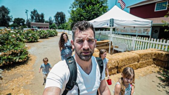 Man with a backpack takes a selfie on a sunny farm path, with kids and a white tent in the background.