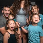 Family portrait in a living room: a smiling dad and mom with four kids gathered around the mom's pregnant belly, celebrating the upcoming baby.