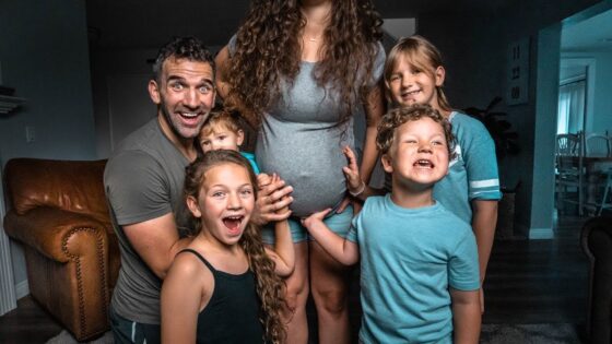Family portrait in a living room: a smiling dad and mom with four kids gathered around the mom's pregnant belly, celebrating the upcoming baby.