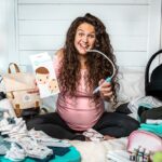 Pregnant woman sitting cross‑legged on a bed, smiling and using a breast pump while surrounded by baby items and a diaper bag in a cluttered nursery setup.