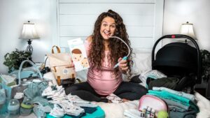 Pregnant woman sitting cross‑legged on a bed, smiling and using a breast pump while surrounded by baby items and a diaper bag in a cluttered nursery setup.