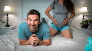 Smiling man lying on a bed with a pregnant woman kneeling behind him, hands on her baby bump in a cozy, lamp-lit bedroom.