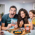 Family of four at a bright kitchen island, smiling as they show snack packets and a pineapple on the counter.