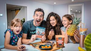 Family of four at a bright kitchen island, smiling as they show snack packets and a pineapple on the counter.