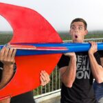Three men on a balcony hold a oversized red and blue prop with surprised expressions on their faces.