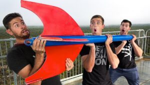 Three men on a balcony hold a oversized red and blue prop with surprised expressions on their faces.