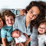 Mother and five children cuddled together on a bed, all smiling at the camera with a newborn infant held by the mother.