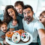 Family selfie with parents and kids gathered around a plate of colorful donuts, smiling in a bright home kitchen/room.