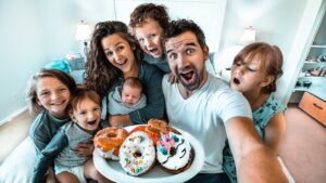 Family selfie with parents and kids gathered around a plate of colorful donuts, smiling in a bright home kitchen/room.