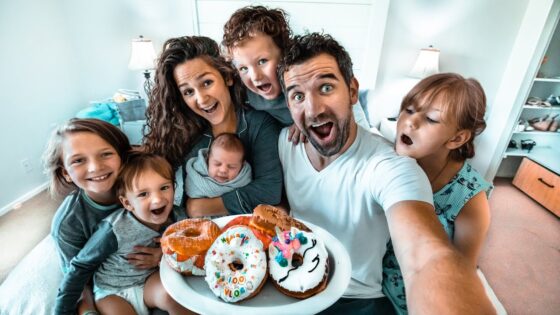 Family selfie with parents and kids gathered around a plate of colorful donuts, smiling in a bright home kitchen/room.