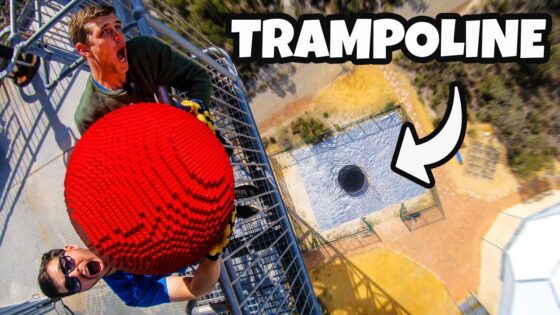 Two people on a tall metal scaffold holding a large bright red spiked ball, with an aerial view of a platform with a hole below.