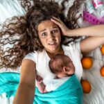 Top-down view of a stressed mother lying on a white quilt with a newborn resting on her chest, wrapped in a turquoise blanket, orange fruits scattered nearby, and baby items in the corner.