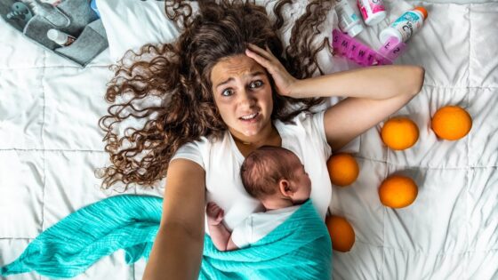 Top-down view of a stressed mother lying on a white quilt with a newborn resting on her chest, wrapped in a turquoise blanket, orange fruits scattered nearby, and baby items in the corner.