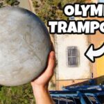 Aerial view of a person holding a large gray sphere above a trampoline setup on a tall structure, with a bold 'OLYMPIC TRAMPOLINE' caption.