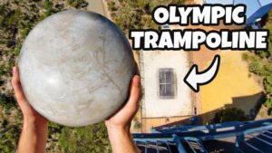 Aerial view of a person holding a large gray sphere above a trampoline setup on a tall structure, with a bold 'OLYMPIC TRAMPOLINE' caption.