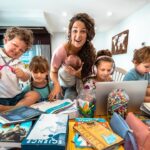 Mother with several children doing homework at a cluttered home table, surrounded by books, crayons, and a laptop in a cozy learning space.
