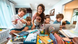 Mother with several children doing homework at a cluttered home table, surrounded by books, crayons, and a laptop in a cozy learning space.