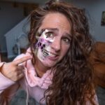 Woman in a pink hoodie points to a half-skull prosthetic makeup on her face, with purple nerve-like lines, in a living room setting behind a brown sofa.