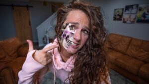 Woman in a pink hoodie points to a half-skull prosthetic makeup on her face, with purple nerve-like lines, in a living room setting behind a brown sofa.