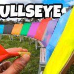 Hand holding a red dart ready to throw at a rainbow-colored target row outdoors, with 'BULLSEYE' text overlaid on the image.