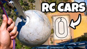 From above: a crane hook grips a weathered gray ball near a small RC car race track, with a bold 'RC CARS' overlay.