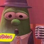 Green VeggieTales character in a hat and tie singing into a vintage microphone on a pink stage with VeggieTales branding nearby.