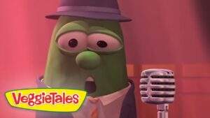 Green VeggieTales character in a hat and tie singing into a vintage microphone on a pink stage with VeggieTales branding nearby.
