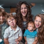 Smiling mom with four kids posing indoors; two children hold scissors as if giving a playful haircut in a cozy living room.
