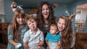 Smiling mom with four kids posing indoors; two children hold scissors as if giving a playful haircut in a cozy living room.