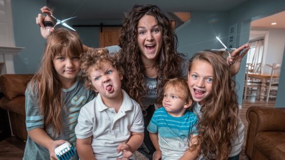 Smiling mom with four kids posing indoors; two children hold scissors as if giving a playful haircut in a cozy living room.