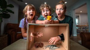 Three children lean over a cardboard box, mouths open in shock as a rubber snake and toy figures lie inside the box at their feet.