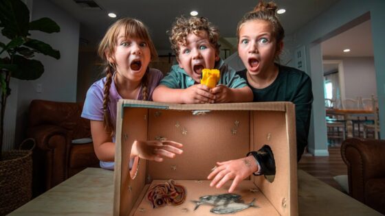 Three children lean over a cardboard box, mouths open in shock as a rubber snake and toy figures lie inside the box at their feet.