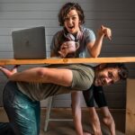 Woman at a DIY desk with laptop and a mug, pointing excitedly as a man crawls under the desk smiling.