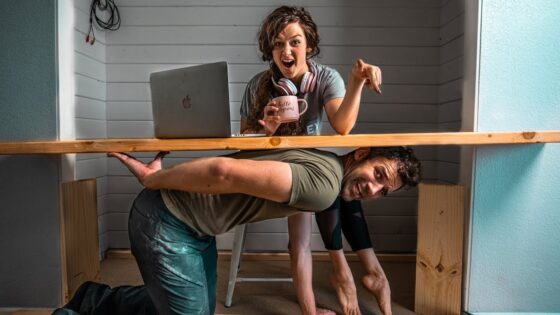 Woman at a DIY desk with laptop and a mug, pointing excitedly as a man crawls under the desk smiling.