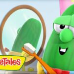Green VeggieTales cucumber character brushing teeth with an orange toothbrush in front of a round mirror in a bright bathroom.