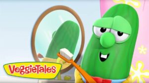 Green VeggieTales cucumber character brushing teeth with an orange toothbrush in front of a round mirror in a bright bathroom.