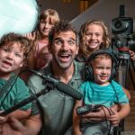 Man and four children posing with a home video setup: camera on a tripod, boom mic, headphones, and a large soft light panel nearby.