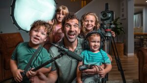 Man and four children posing with a home video setup: camera on a tripod, boom mic, headphones, and a large soft light panel nearby.