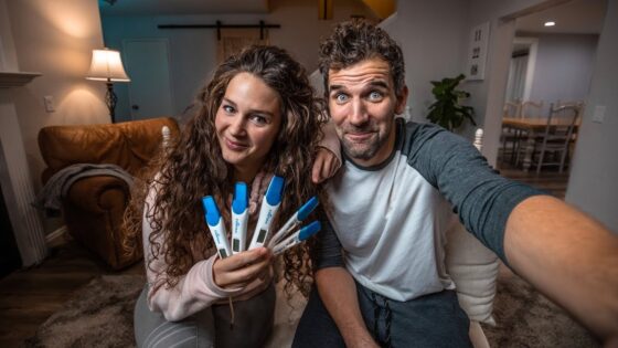Couple sitting on a living room floor, smiling, holding several blue-cipped pregnancy tests toward the camera.