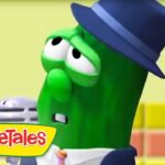 Green VeggieTales character wearing a blue hat, holding a microphone, on a colorful set with the logo in the corner.