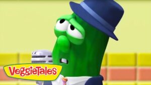Green VeggieTales character wearing a blue hat, holding a microphone, on a colorful set with the logo in the corner.