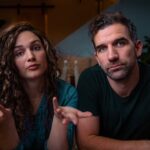 Two adults sit on a couch in a dimly lit living room; the woman gestures with open hands as she speaks, while the man looks at the camera.