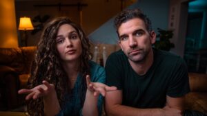 Two adults sit on a couch in a dimly lit living room; the woman gestures with open hands as she speaks, while the man looks at the camera.