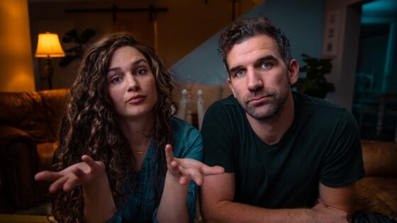 Two adults sit on a couch in a dimly lit living room; the woman gestures with open hands as she speaks, while the man looks at the camera.