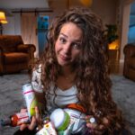 Woman with long curly hair sitting on a living room rug, holding a pile of shampoos and toiletries.