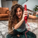 Woman sitting on a living room rug, using a red hair styling tool on her long curly hair with a playful, intense expression.