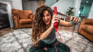 Woman sitting on a living room rug, using a red hair styling tool on her long curly hair with a playful, intense expression.