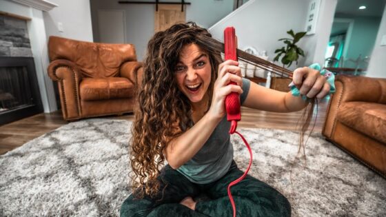 Woman sitting on a living room rug, using a red hair styling tool on her long curly hair with a playful, intense expression.