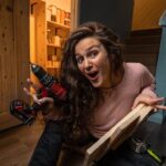 Woman kneeling on a workshop floor holds a cordless drill and a wooden plank with a surprised expression, surrounded by tools and wood.
